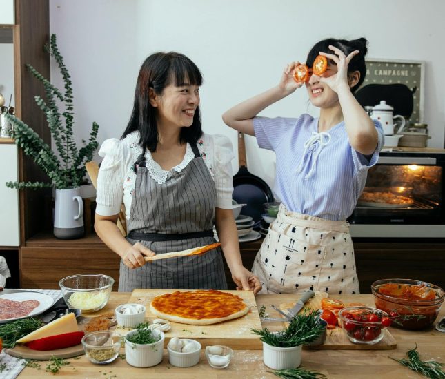 Two friends joyfully make pizza together in a bright, inviting kitchen filled with fresh ingredients.